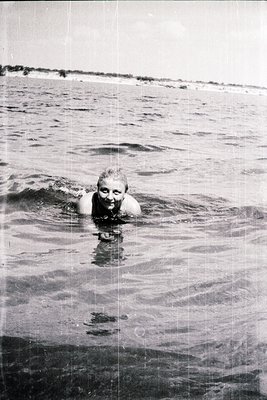 Vintage black-and-white photo of a woman swimming in calm ocean waters, mid-20th century beachwear. Gentle waves and distant ...