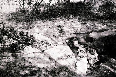 Black-and-white photo of a person lying prone on a sandy, barren landscape with scattered debris. Clothing appears worn and t...
