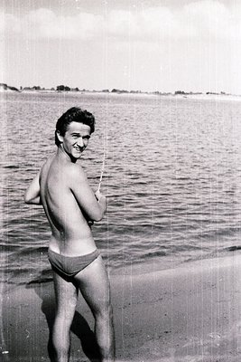Young man in mid-20th-century swim trunks poses by a serene coastal shoreline, holding a fishing rod. Black-and-white vintage...