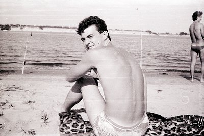 Mid-20th century beach scene featuring a shirtless man in patterned swim trunks, seated on a towel. Soft waves and shallow wa...
