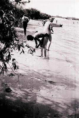Black-and-white beach scene featuring three shirtless men in mid-1960s swimwear, one bending to pick up a shell near shallow ...
