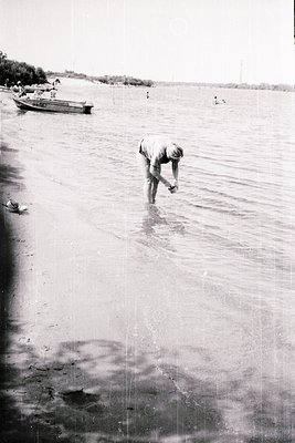 Mid-20th century black-and-white seaside scene: lone fisherman wading in shallow water, holding a net or line. Small wooden b...