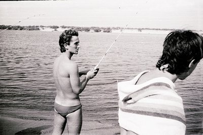 Black-and-white shot of two men fishing at a serene lakeside, mid-20th century. One man in swim trunks casts a line; the othe...