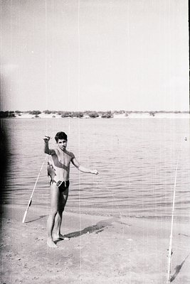 Vintage black-and-white seaside shot of a young man in minimalist swimwear posing with a long pole, likely for windsurfing. S...