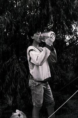 Black-and-white candid of a person drinking from a glass bottle under willow branches, mid-20th century. Casual attire—rolled...