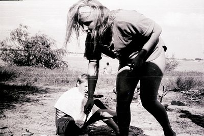 Black-and-white photo capturing a moment of care in a rural setting. A woman assists a seated child, likely tending to a mino...