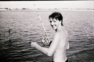 Shallow-water fishing scene with man holding a spinning reel and freshly caught fish, mid-1970s. Classic vintage black-and-wh...