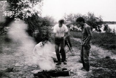 Vintage black-and-white photo of three individuals gathered around an outdoor fire in a wooded area, likely mid-20th century....