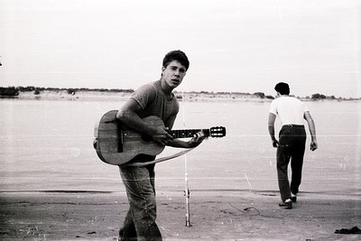 Young man in 1960s-70s beachwear holds acoustic guitar on sandy shore, mid-coastal walk. Second figure in white shirt walks a...