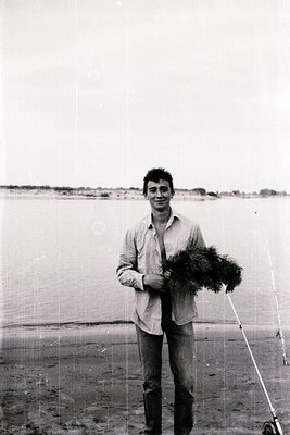 Black-and-white portrait of a young man holding a bundle of reeds by a shallow, reflective body of water, likely a lake or co...