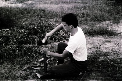 Man in mid-20th-century outdoor setting, squatting beside a small metal structure with a bucket, possibly filling or emptying...