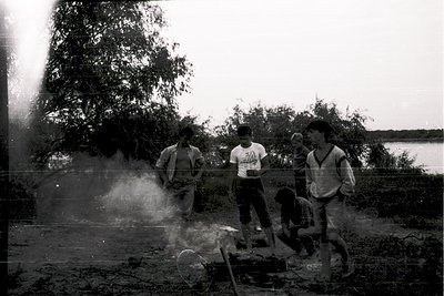 Black-and-white photo of four young men by a waterfront, likely mid-20th century. One man grills food over an open fire, whil...