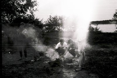 Vintage black-and-white photo of three men tending a small fire in an outdoor setting, likely rural. Steam rises from a metal...