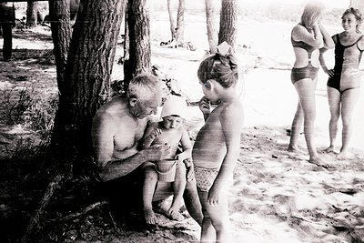 Mid-20th century beach scene: shirtless man in swim trunks hugs a child while another child stands beside him. Two women in s...