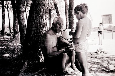 Mid-20th century seaside scene: an elderly man in swim trunks holds a child in a striped swimsuit, feeding them from a white ...