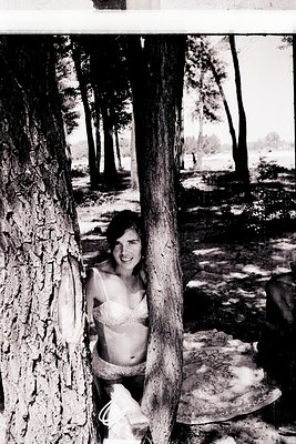 Black-and-white candid of a woman peeking from behind a tree trunk, wearing a lace bikini top and patterned bottoms. Natural ...