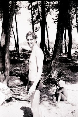 Young man in swim trunks poses confidently in a forested lakeside setting, mid-20th century. Trees frame the scene, with anot...