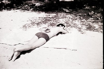 Black-and-white shot of a man reclining on a sandy beach, mid-20th century. Wearing patterned swim trunks, he rests on his st...
