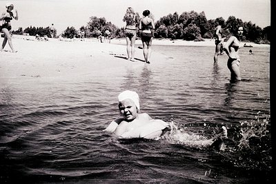 Mid-20th century beach scene featuring a child in a full-body swim diaper wading in shallow water. Surrounding adults and chi...