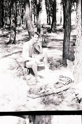 Vintage black-and-white photo of a shirtless man seated on a tree stump in a wooded area, resting his chin on his hand. Folde...