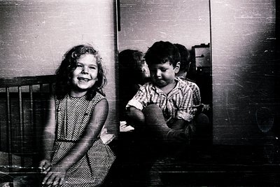 Two children in a mid-20th-century indoor setting, likely 1950s–1960s. Girl in patterned dress leans on a metal-framed bed, s...