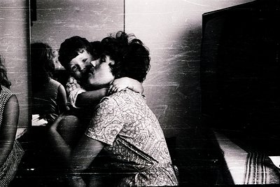 Intimate black-and-white moment of two children hugging in a dimly lit indoor setting, likely a home. The woman’s patterned b...