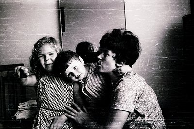 Vintage black-and-white photo of three children hugging indoors, likely mid-20th century. The older girl points at the camera...