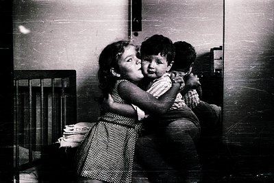 Two children hugging indoors, mid-20th century. Girl in patterned dress clings to boy in striped shirt; third child watches f...