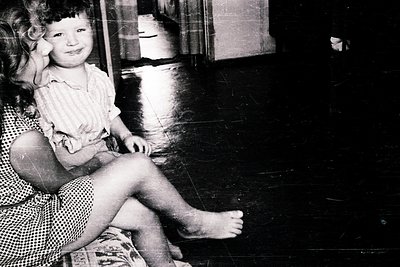 Black-and-white portrait of a young girl (approx. 4-6 years) seated on a woman’s lap in a dimly lit room. The girl wears a st...