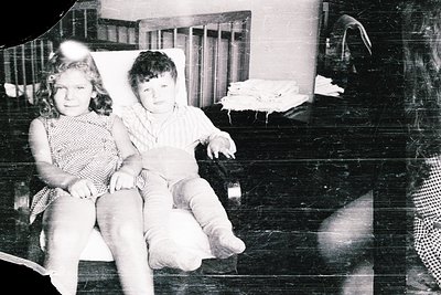 Two children pose indoors on a sofa in a mid-20th-century setting. Girl in a sleeveless dress with a patterned bodice, boy in...