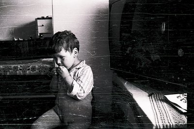 Mid-20th century black-and-white photo of a young boy in a modest indoor setting, likely residential. He stands near a tiled ...