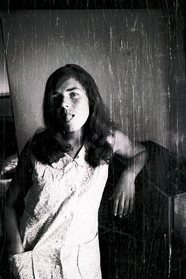 Vintage black-and-white portrait of a woman in a 1960s-style lace blouse, leaning against a textured wall. Her pose and attir...