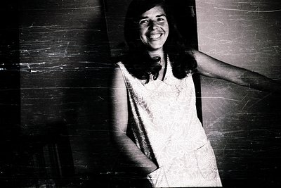 Vintage black-and-white portrait of a woman in a sleeveless lace dress, leaning against a textured wall. Her joyful expressio...
