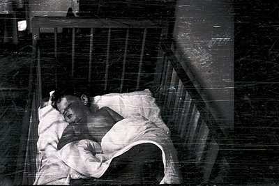 A man rests on a narrow metal-framed cot in a dimly lit, institutional setting. The cot’s thin mattress and worn sheets sugge...