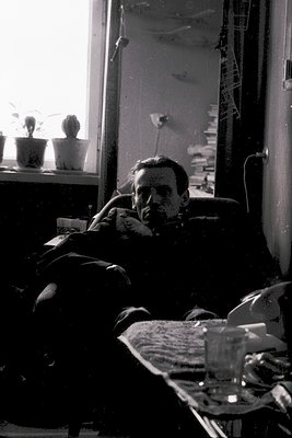 Mid-20th century interior shot: man reclines on a worn sofa beside a glass-topped table with a wineglass. Sunlight streams th...