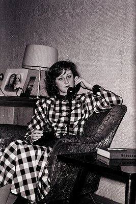 Vintage black-and-white portrait of a woman in a plaid dress, seated on a tufted chair while speaking on a rotary phone. Side...