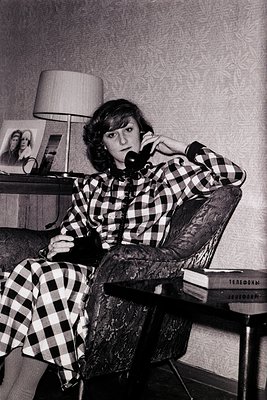 Vintage black-and-white portrait of a woman in a plaid blouse and leather jacket, seated in a mid-century armchair while spea...
