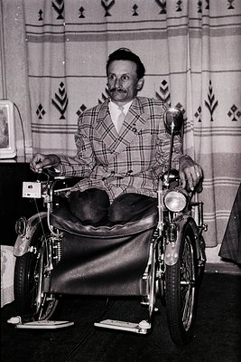 Mid-20th century man in a plaid suit seated in a sidecar-style wheelchair, likely 1950s–1960s. Patterned wallpaper with geome...