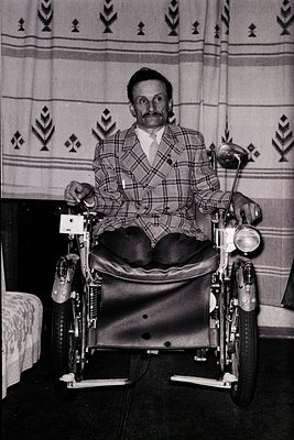 Mid-20th century man in a manual wheelchair, wearing a plaid suit and white shirt, posed indoors against a patterned textile ...