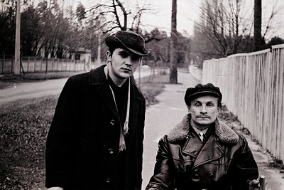 Black-and-white portrait of two men in winter attire—one in a peacoat with scarf, the other in a fur-lined coat and flat cap—...