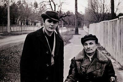 Two men pose outdoors in mid-20th century attire, likely Eastern Bloc era (1950s–1970s). The man on left wears a flat cap, sc...