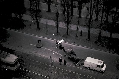 Aerial view of urban street maintenance in mid-20th century. Workers repair a damaged streetlight pole near a traffic circle,...