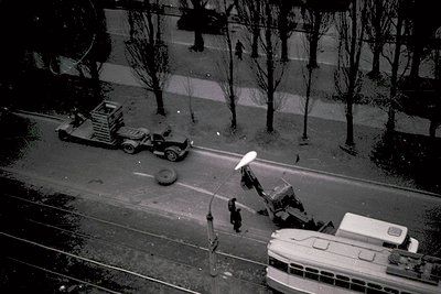 Aerial view of mid-20th century urban roadwork: vintage truck with flatbed hauling debris, parked bus, and construction equip...