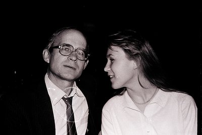 Black-and-white portrait of a man and woman in formal attire, likely from the 1970s–1980s. The man wears round glasses, a whi...