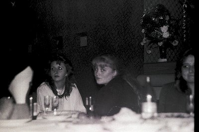 Vintage sepia-toned photo of three women seated at a dimly lit table, likely mid-20th century. Blurred floral wallpaper and w...