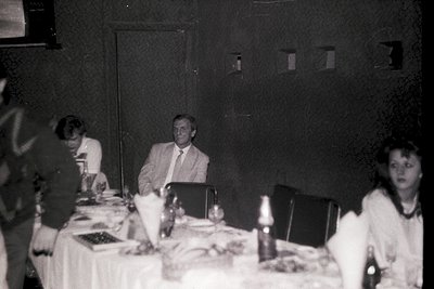 Vintage black-and-white indoor gathering at a formal banquet table, featuring three adults seated. Central figure in suit and...