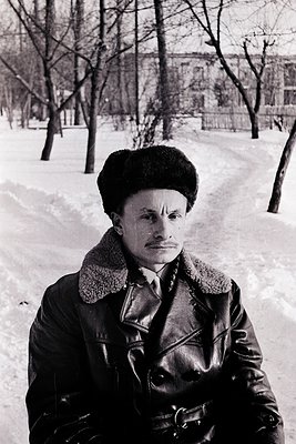 Portrait of a man in a winter coat with fur collar, standing on a snow-covered path in a park. His attire suggests mid-20th c...