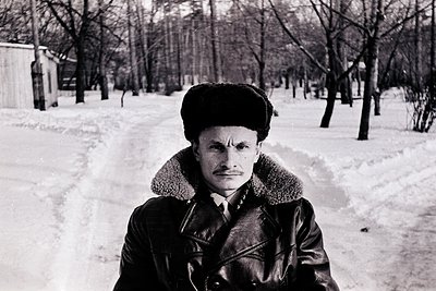 Portrait of a man in winter attire—fur-lined coat, scarf, and fur hat—standing on a snow-covered path in a leafless park. Mid...