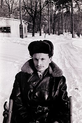 Mid-20th century man in winter attire—fur-lined coat, leather gloves, and fur hat—sitting on a sled in a snow-covered forest....
