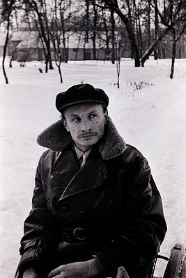 Mid-century man in winter attire—faux-fur collar, leather jacket, and cap—sits on a snow-covered path. Leaves remain on leafl...